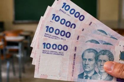 Mano con billetes argentinos de diez mil pesos frente a un aula