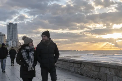 Personas caminan abrigadas junto al mar con algo de sol entre las nubes